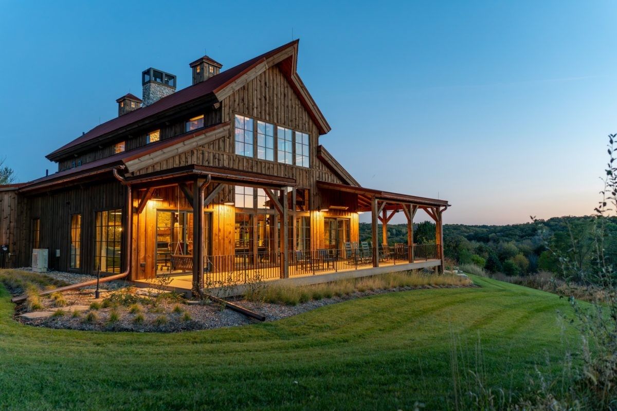 Rustic wooden house with warm porch lights at dusk, set on a grassy hillside under a blue sky.