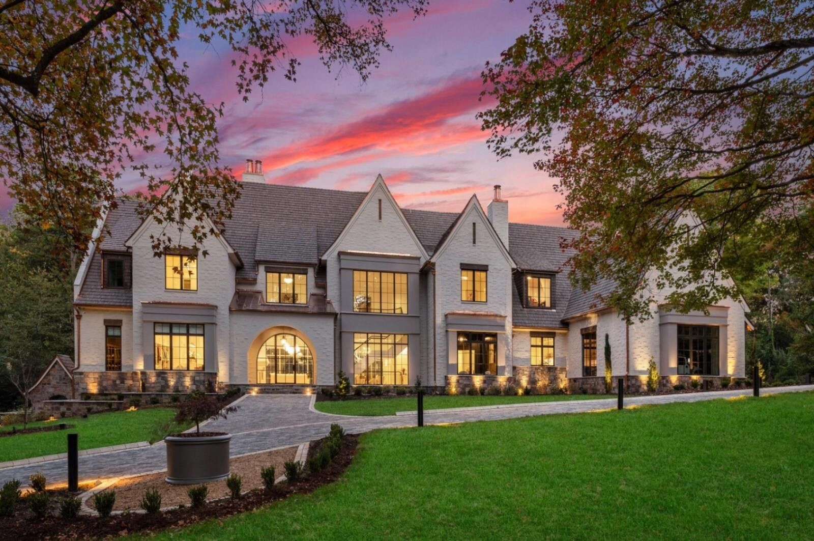 Large gray luxury home at dusk with warm lights, manicured lawn, and pink sunset sky