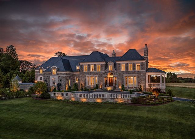 Grand stone mansion at sunset with warm lights and a dramatic pink-orange sky over a manicured lawn