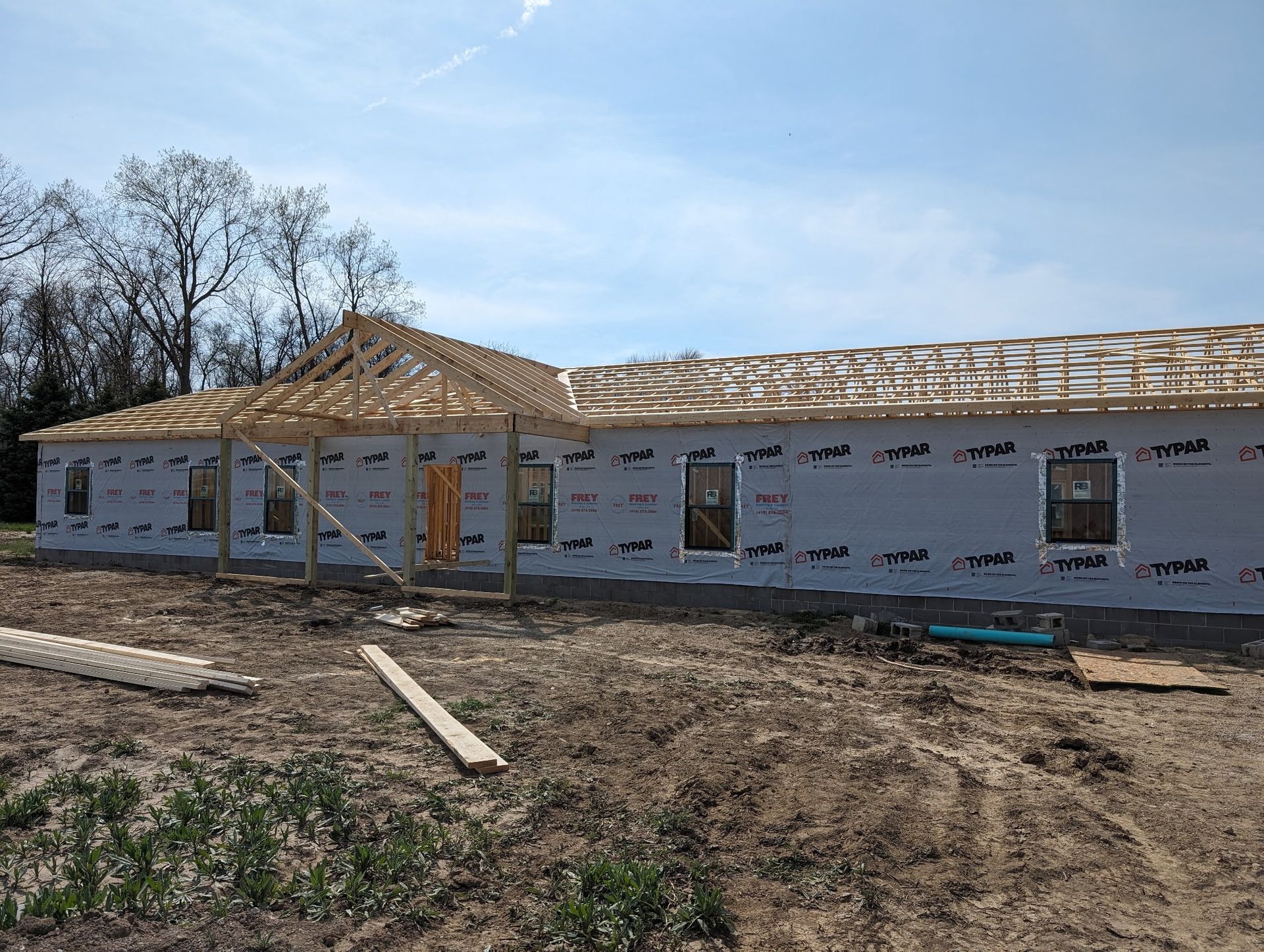 Partially built house with white wrap, exposed roof trusses, and dirt yard under a clear sky