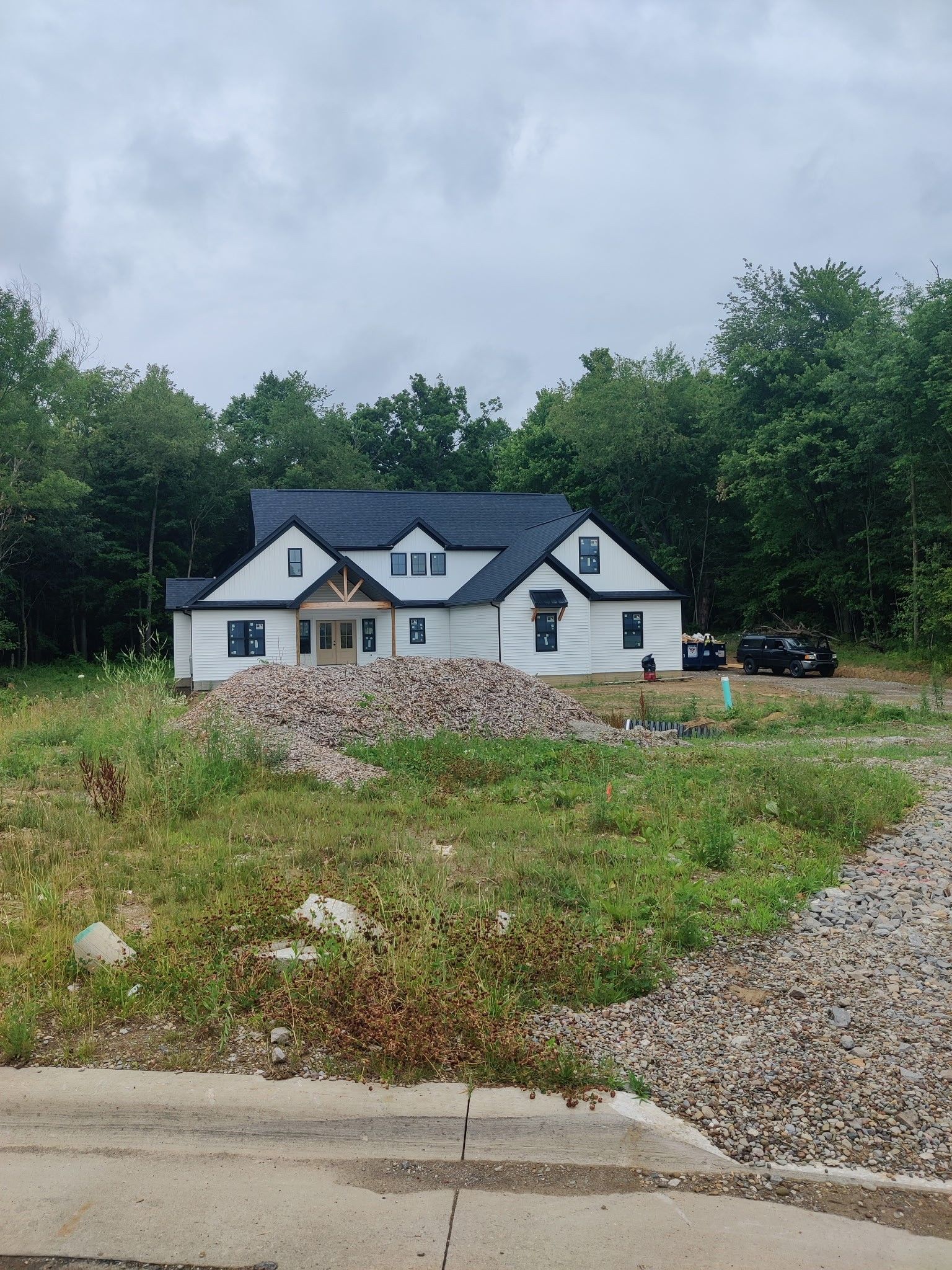 White house under construction on a grassy lot beside gravel driveway, with trees and cloudy sky.