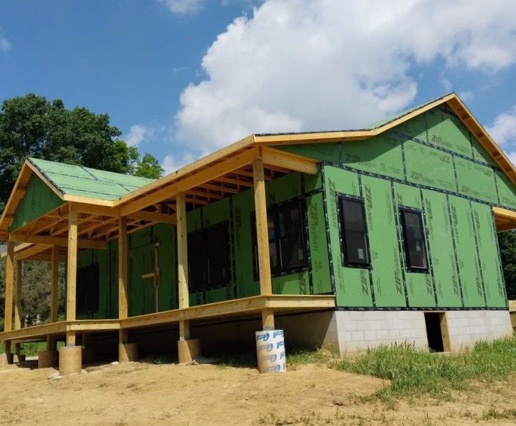 Green house under construction with a wooden porch and roof, set on a dirt lot beneath a blue sky.