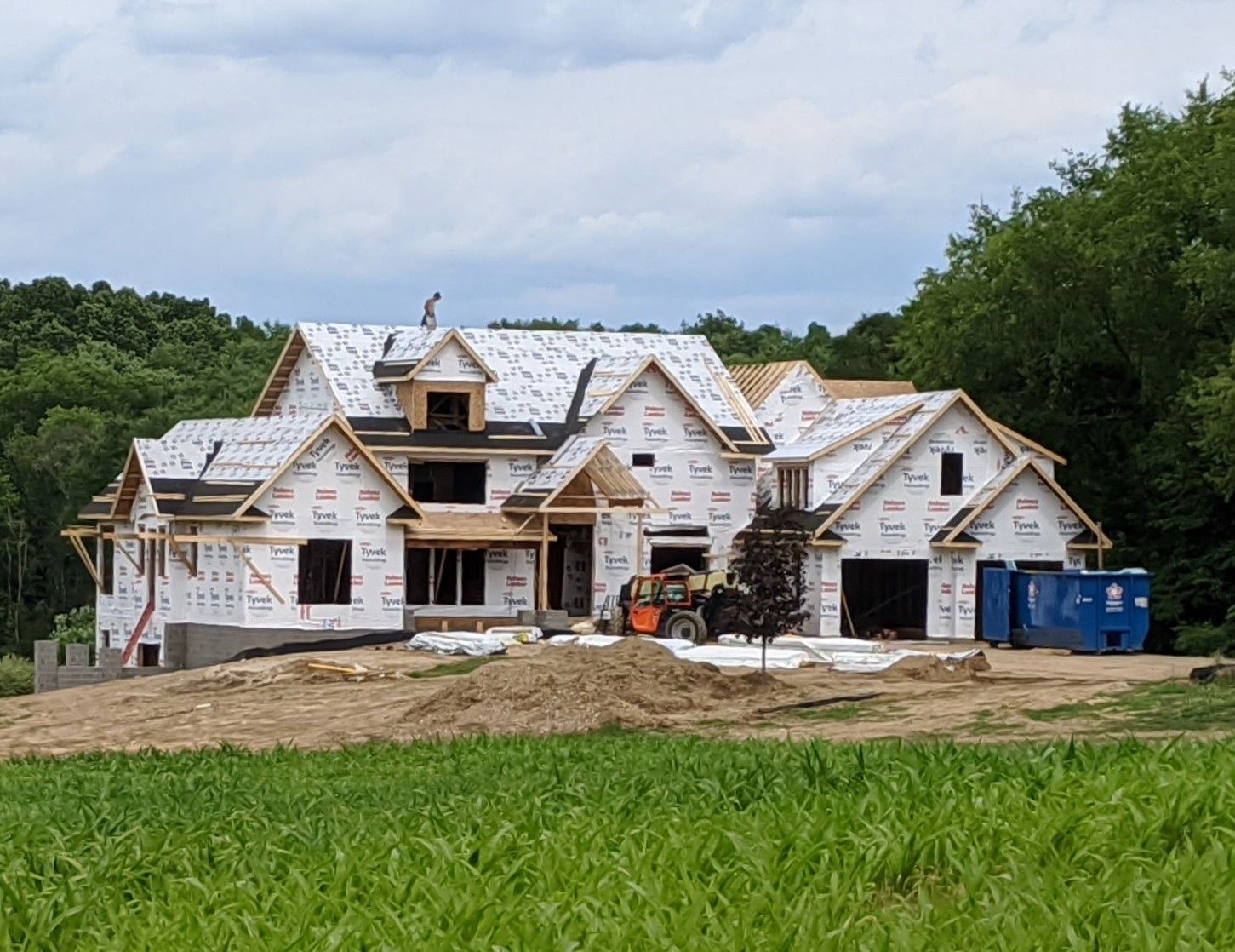 House under construction with white house wrap, dirt foreground, and a blue dumpster beside it