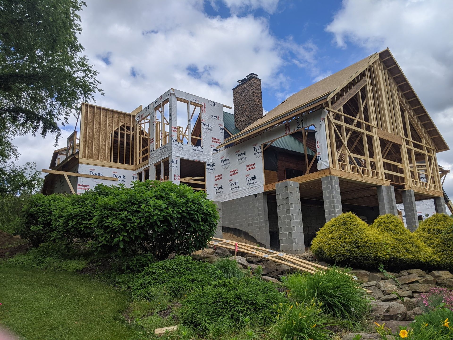 House under construction with exposed wood framing, stone foundation, and blue sky above a grassy slope.