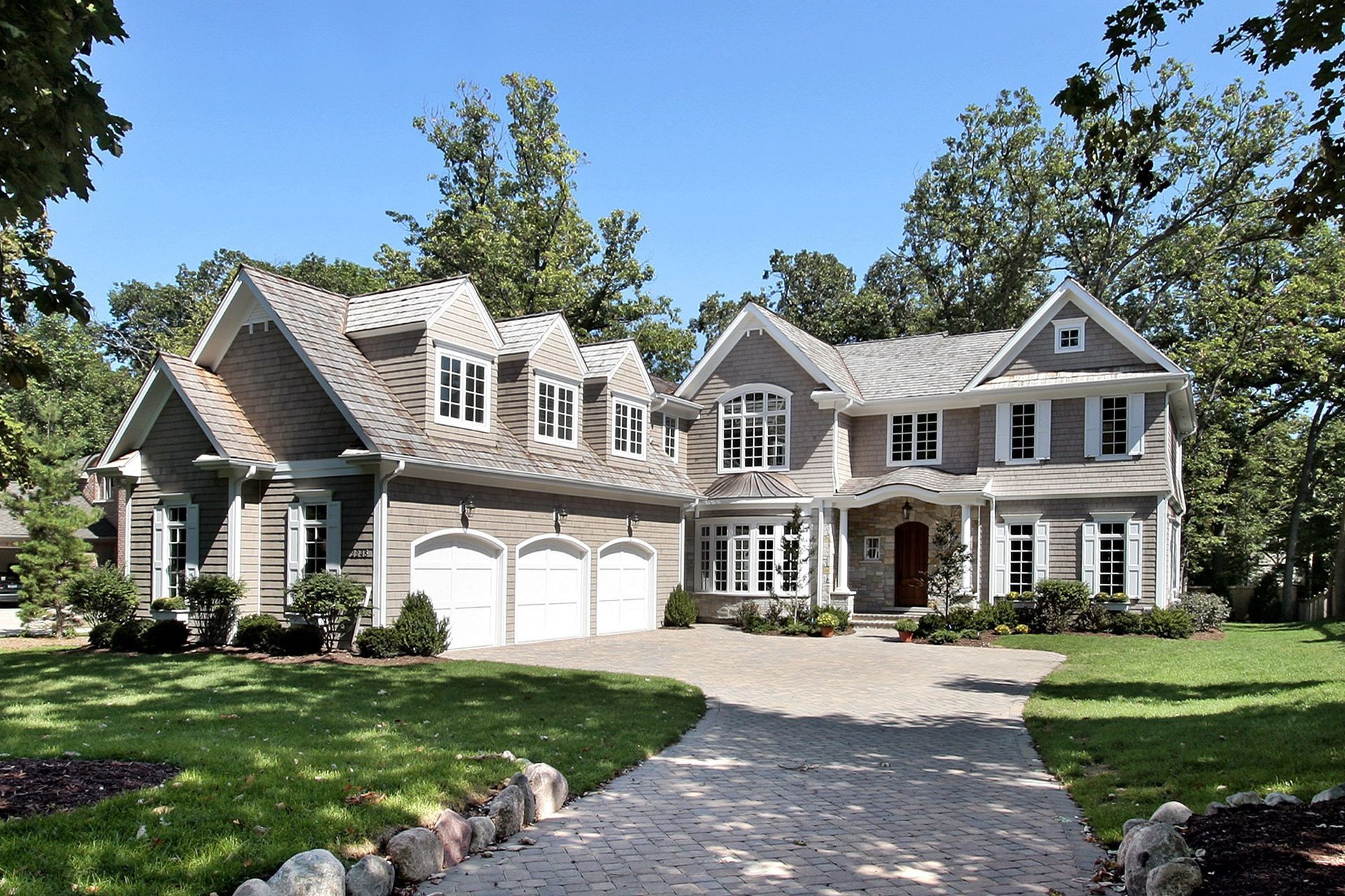 Large gray suburban house with white trim, three-car garage, and curved driveway surrounded by trees