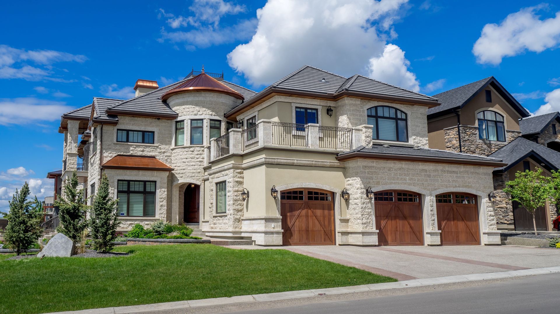 Large stone suburban house with three garage doors, manicured lawn, and blue sky with clouds