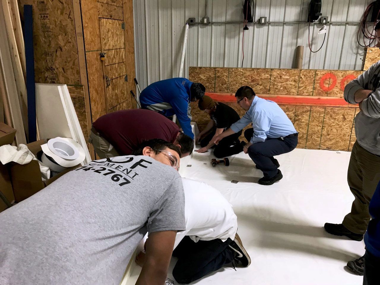 People crouch around a small object on a white floor in a workshop or warehouse.