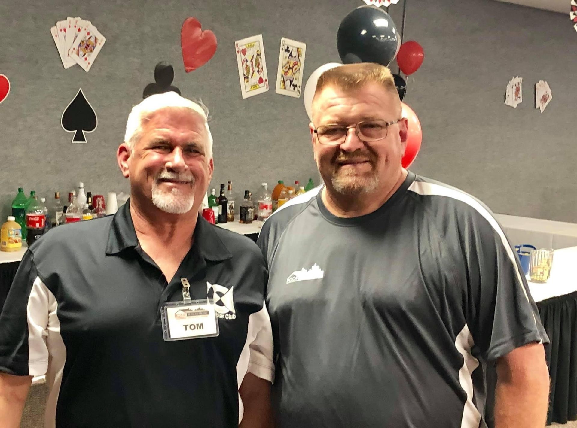 Two smiling men standing indoors at a party, wearing black and gray shirts, with balloons and decorations behind them