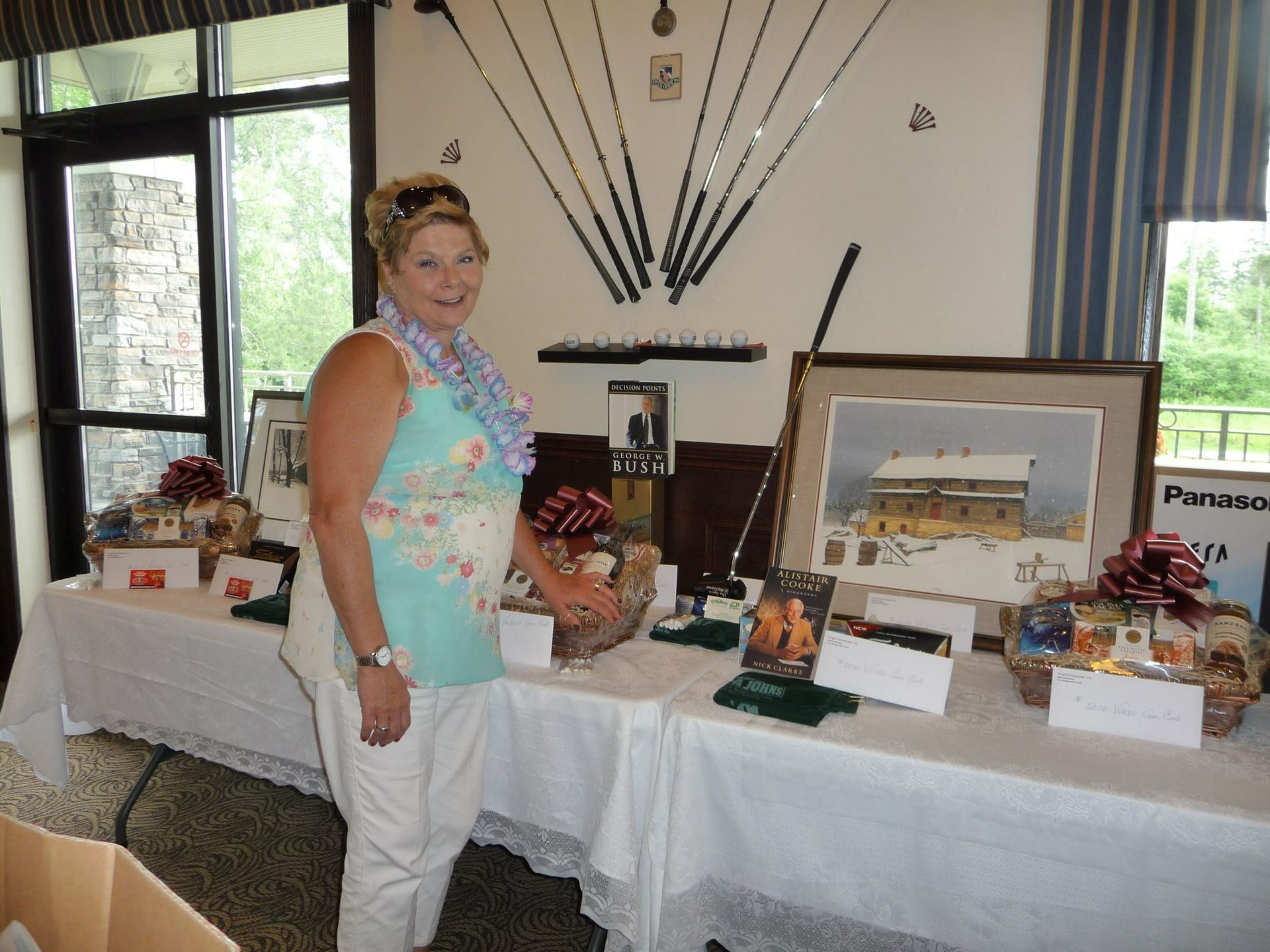 Woman stands behind a display table with framed artwork and decorative items in a bright room.