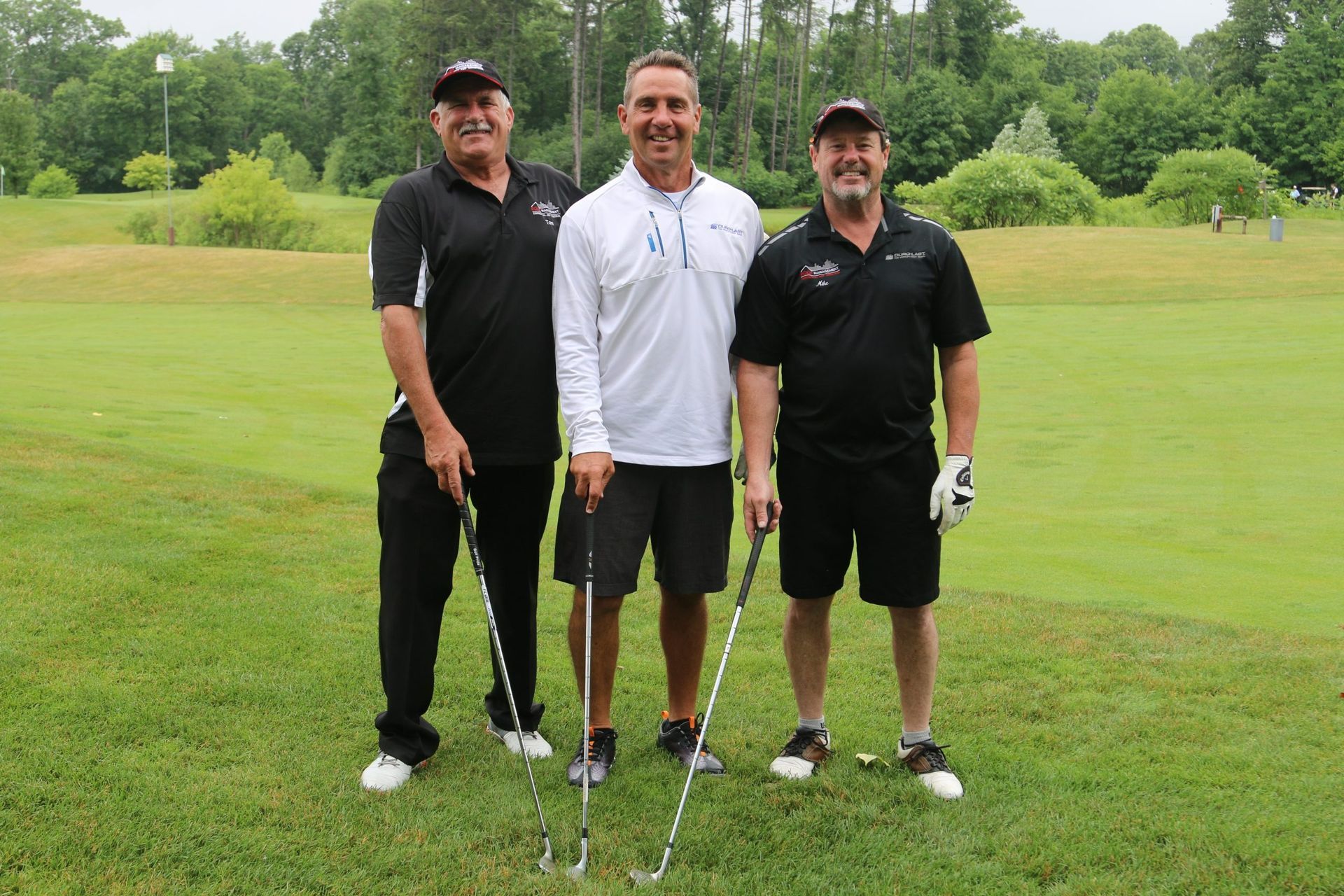 Three golfers standing on a green fairway, holding clubs, with trees in the background.