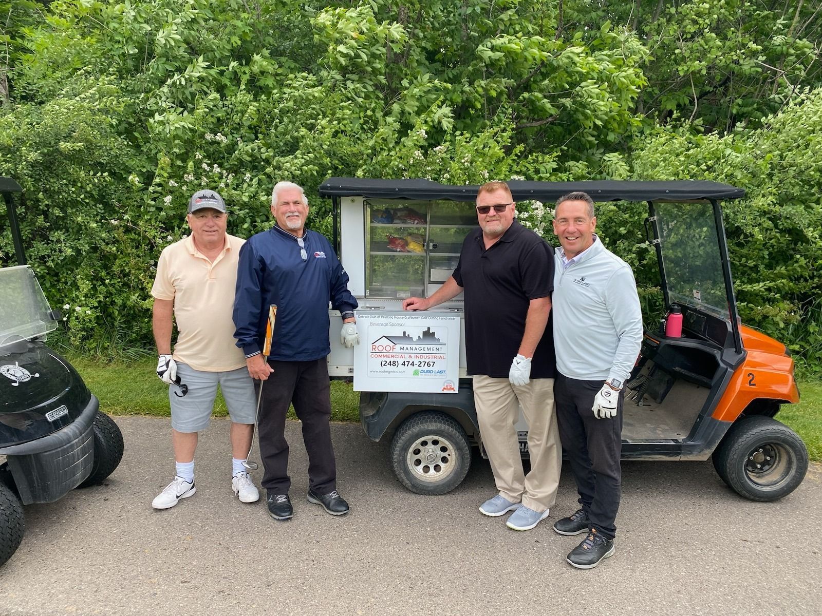 Four people posing beside a small utility vehicle in a wooded outdoor setting, holding a sign.