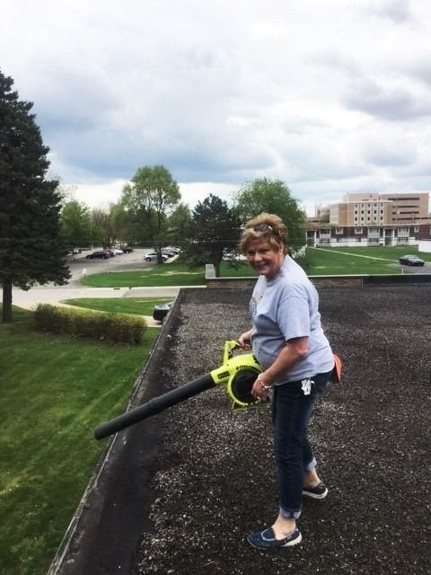 Person using a leaf blower on a rooftop, with grassy courtyard and buildings in the background.