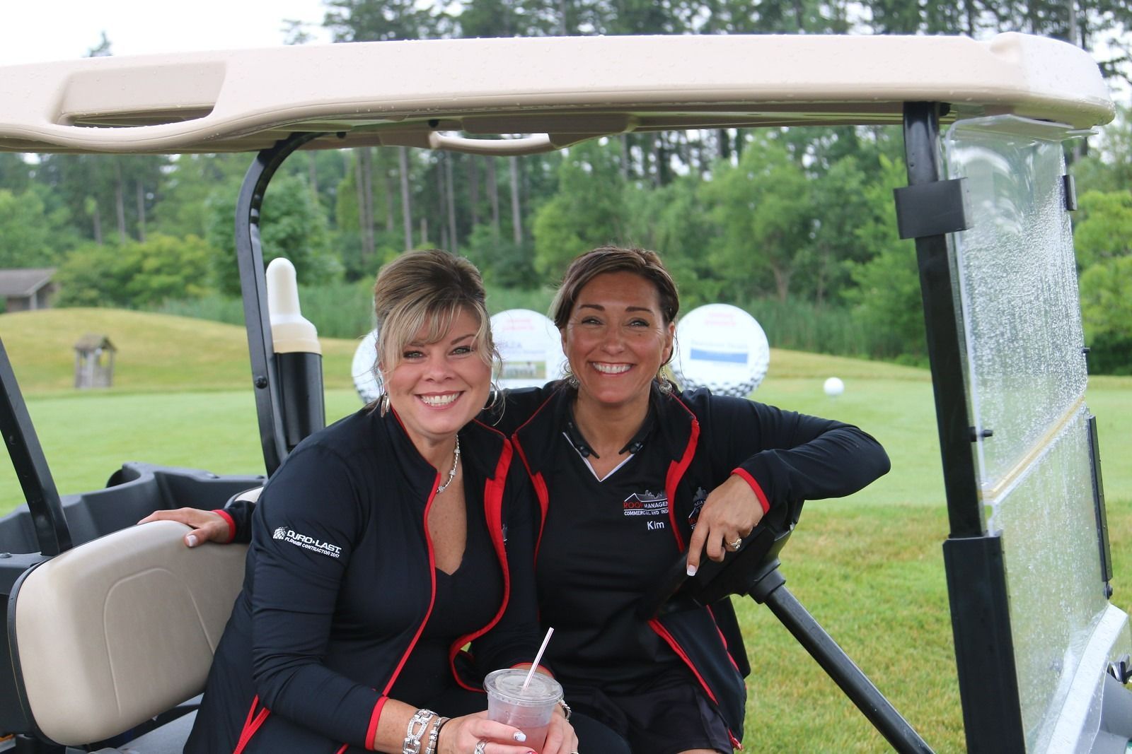 Two people in black-and-red jackets smiling on a golf cart on a grassy course