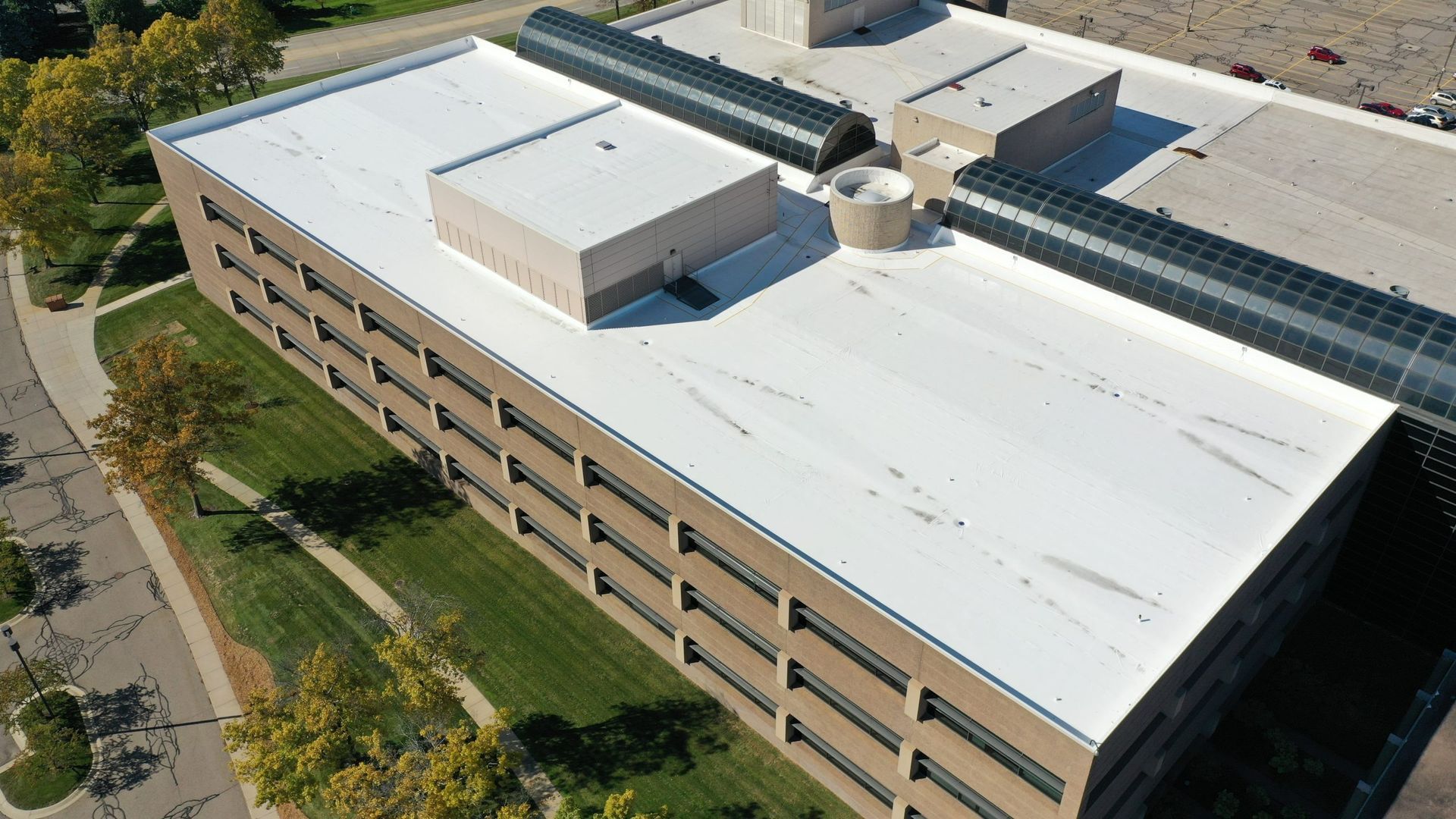 Aerial view of a beige office building with a white roof and rooftop equipment, beside a tree-lined street