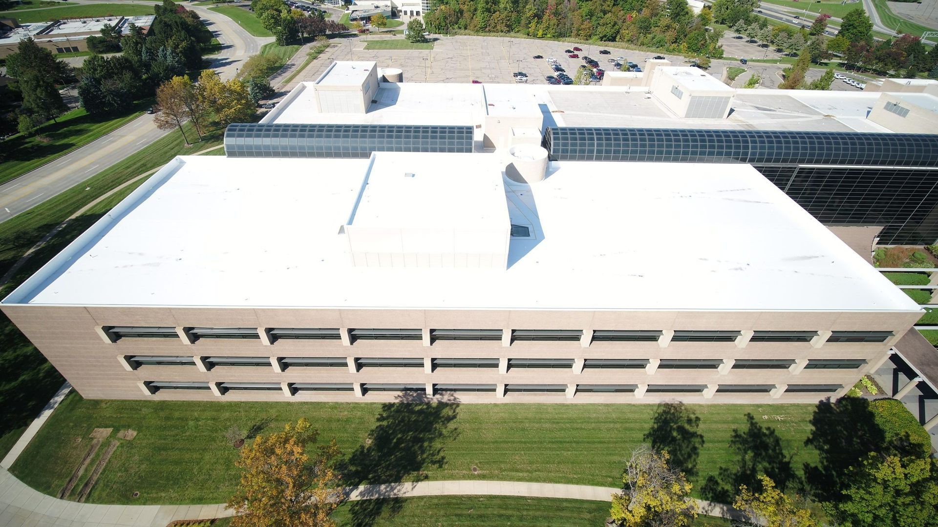 Aerial view of a large beige office building with a white roof and surrounding trees and walkways