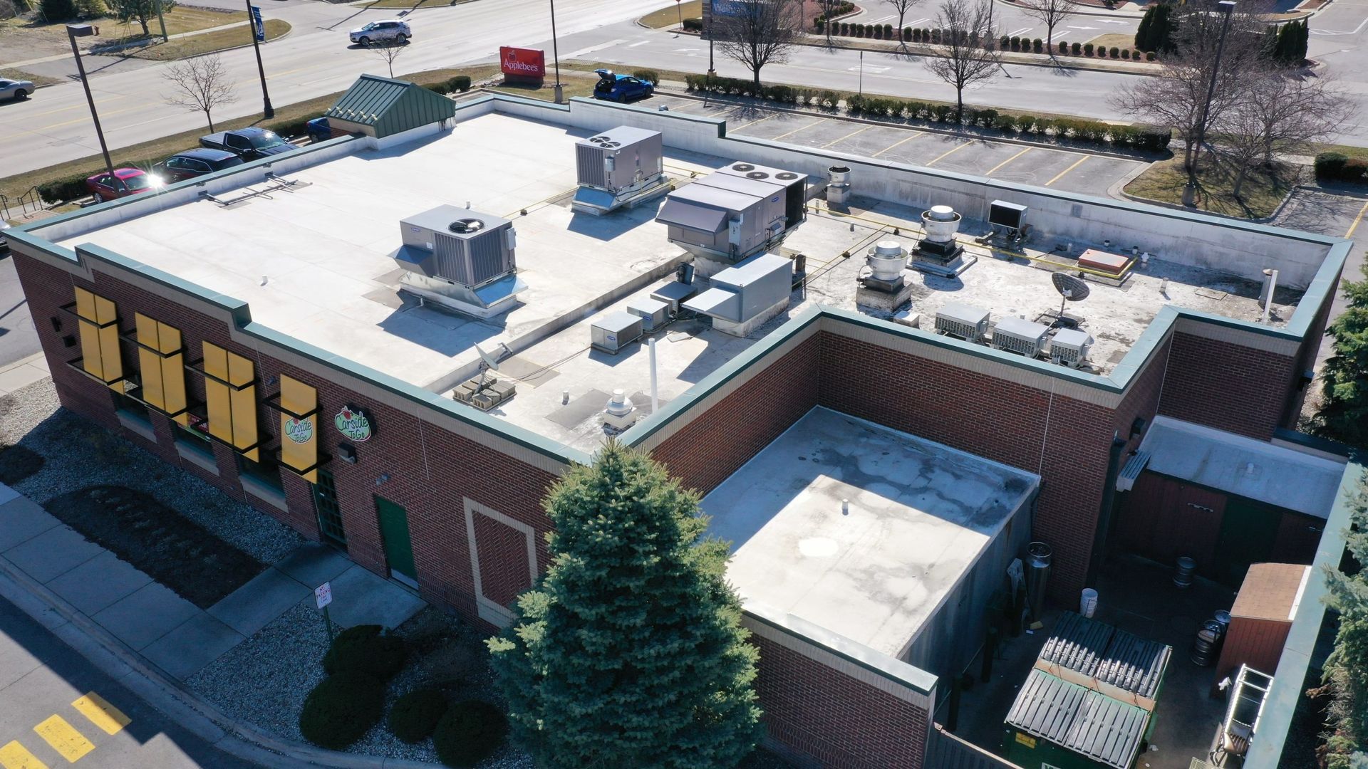 Aerial view of a brick commercial building with a flat white rooftop and HVAC units, beside a parking lot.