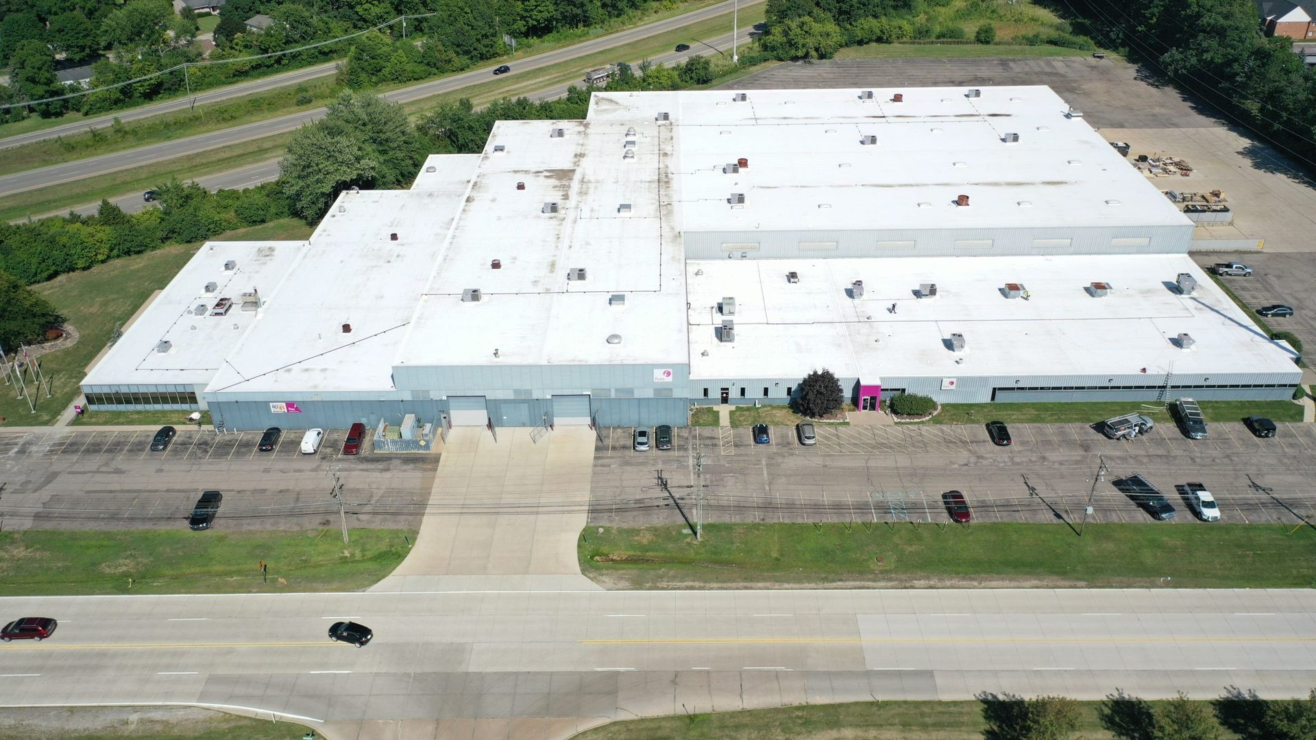 Aerial view of a large white industrial building beside a road and parking lot, with trees and rail lines nearby