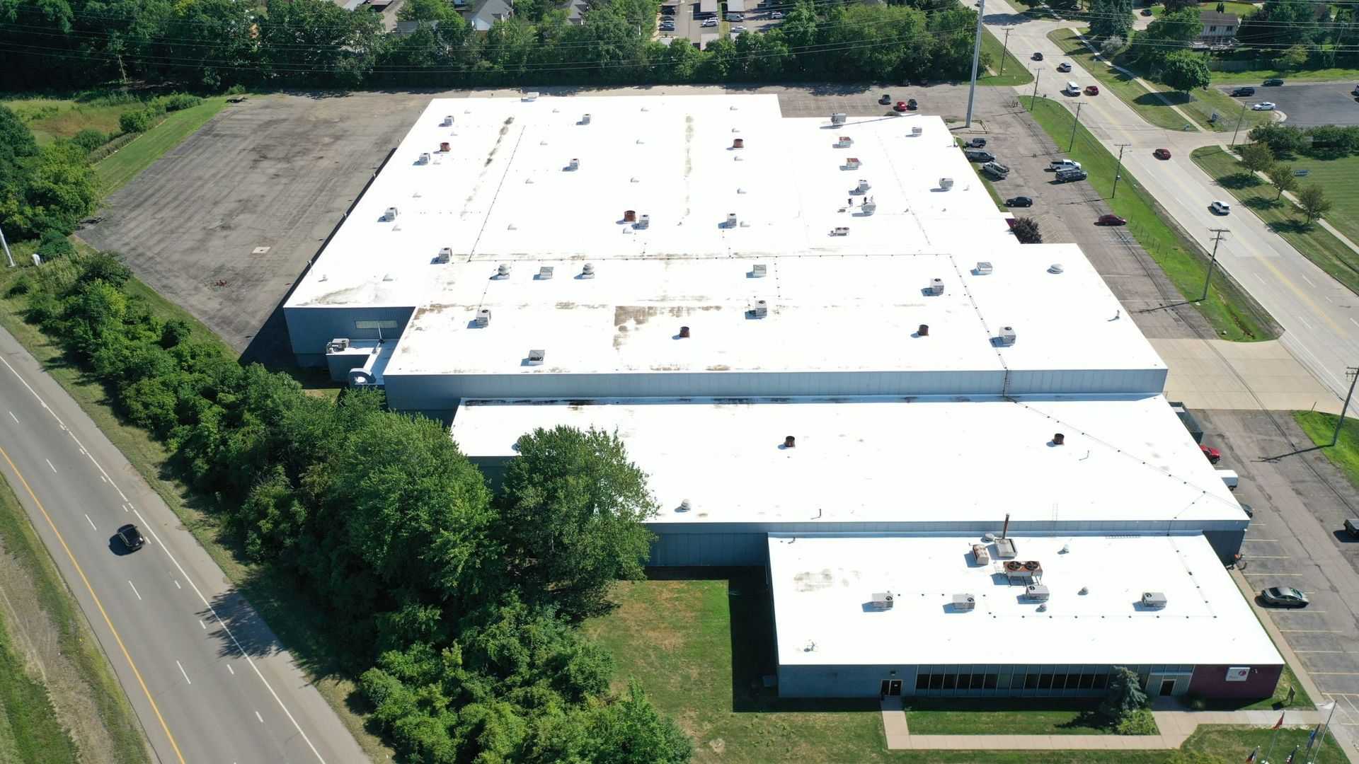 Aerial view of a large white-roofed industrial building beside roads and trees.