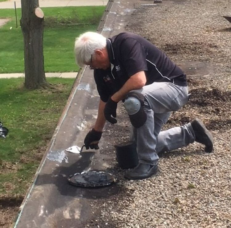 Worker kneeling on gravel, spreading black sealant along a curb with a trowel.