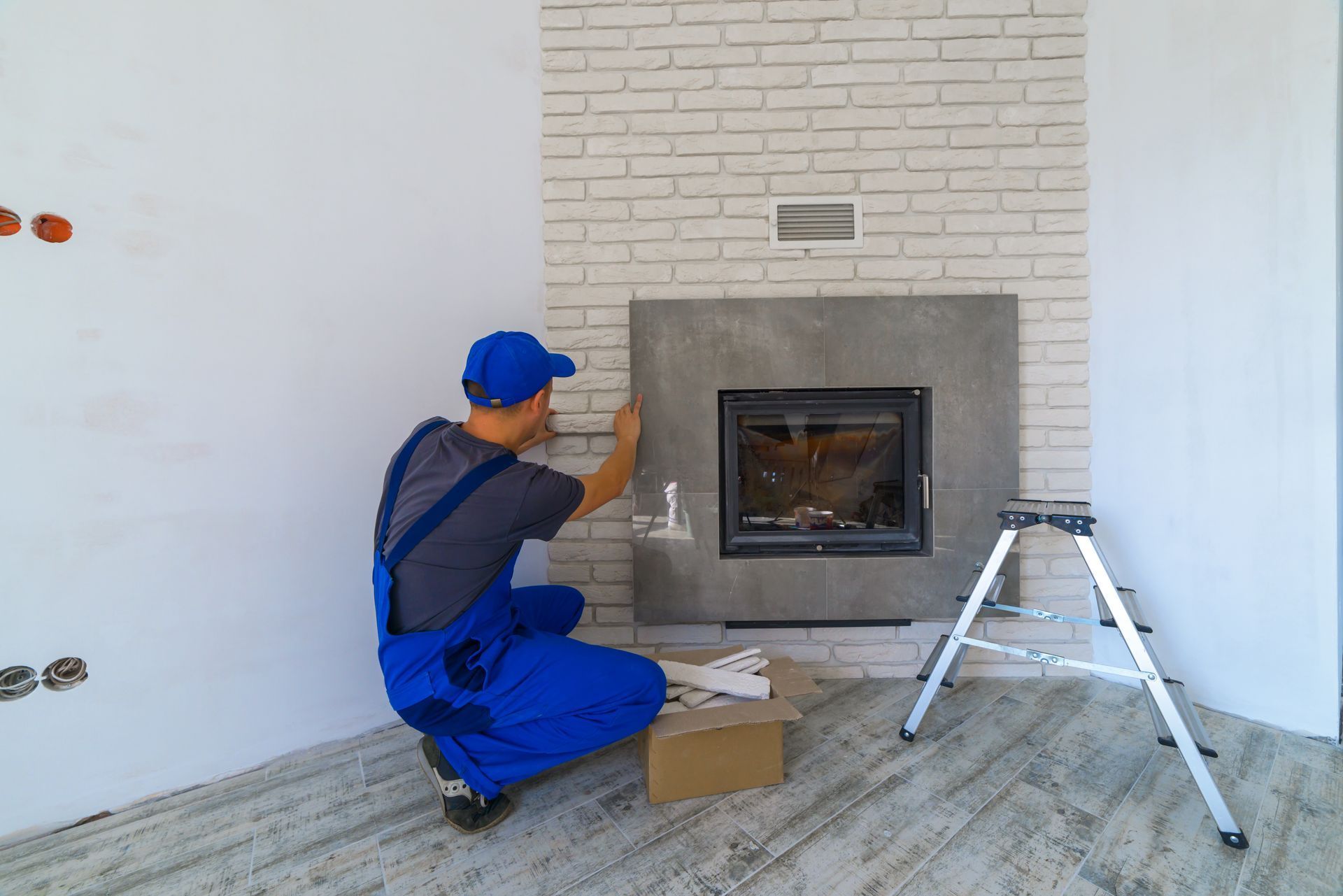 Worker in blue overalls installing a fireplace in a room with a ladder and tools.