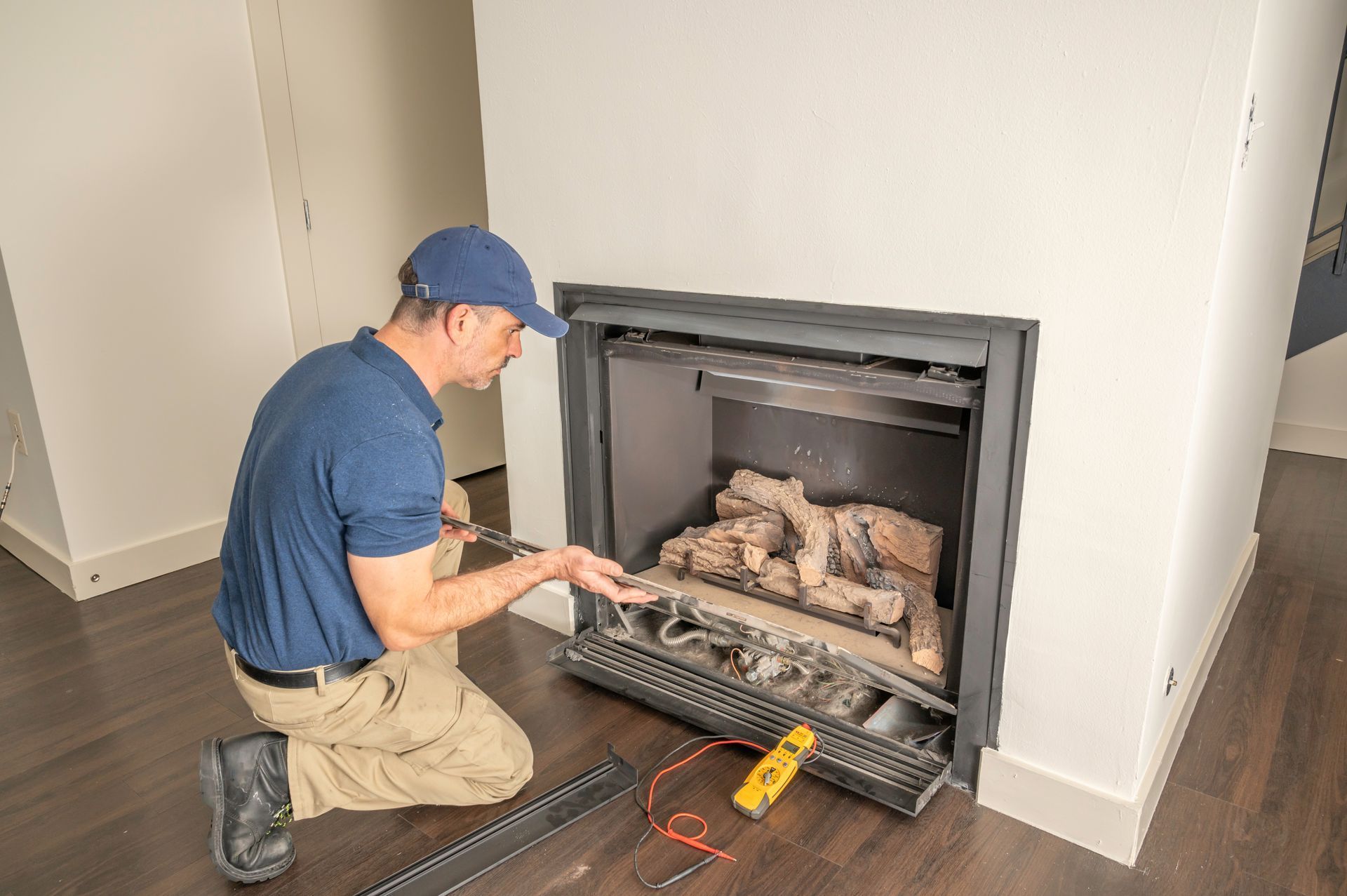 Person kneels by a fireplace, adjusting gas logs with tools in a living room.