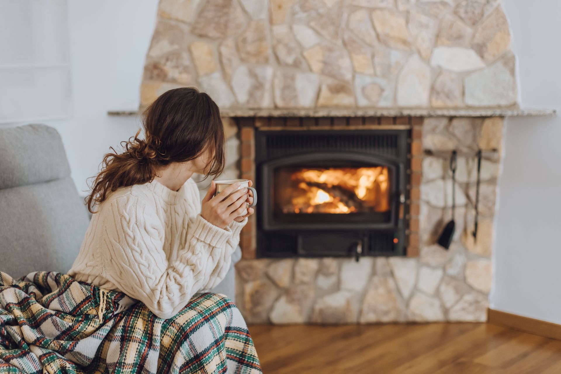 Person wrapped in a blanket sits by a stone fireplace, holding a mug in a cozy living room.