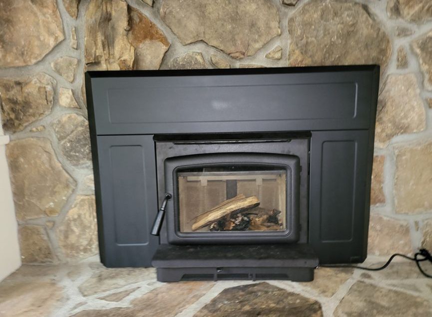 Black wood stove insert set into a stone fireplace, with a glass door and visible logs inside.