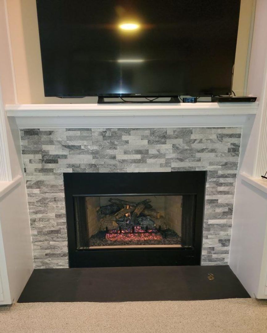 Living room fireplace with gray tile surround, black mantel, and TV above, with a lit fire inside.