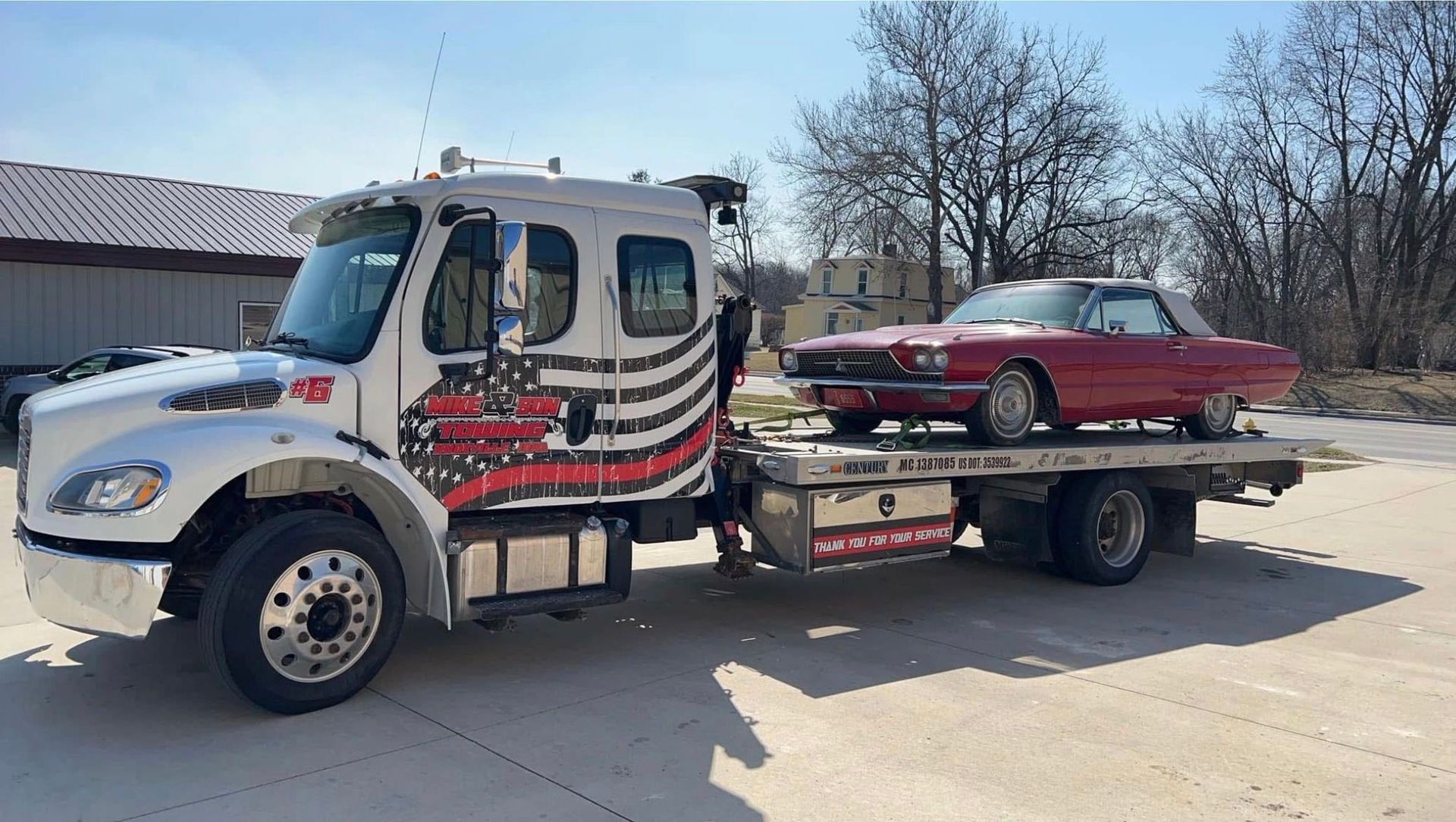 Tow truck carrying a red classic car on a sunny day. The truck is white with red and black graphics.