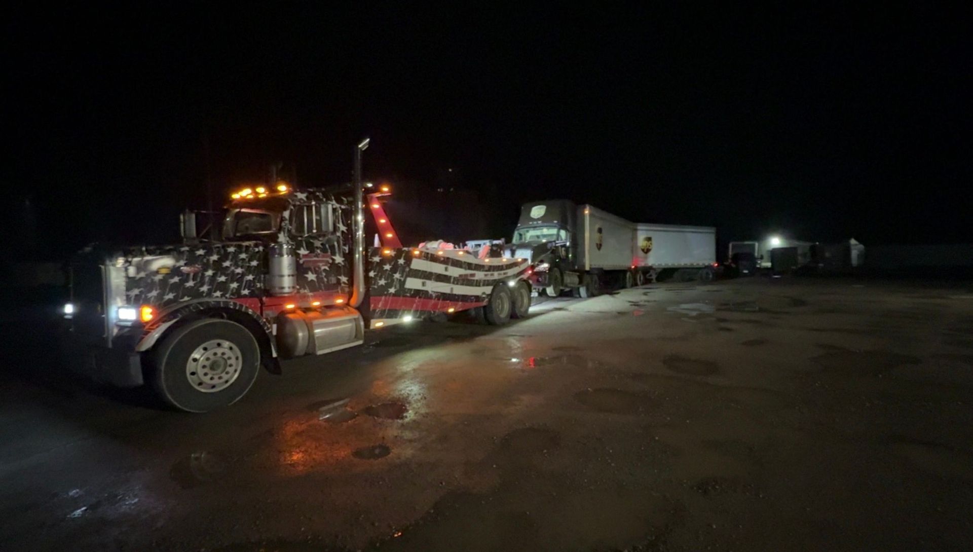 Semi-truck with American flag design pulling a trailer at night. Lights illuminate the vehicles.