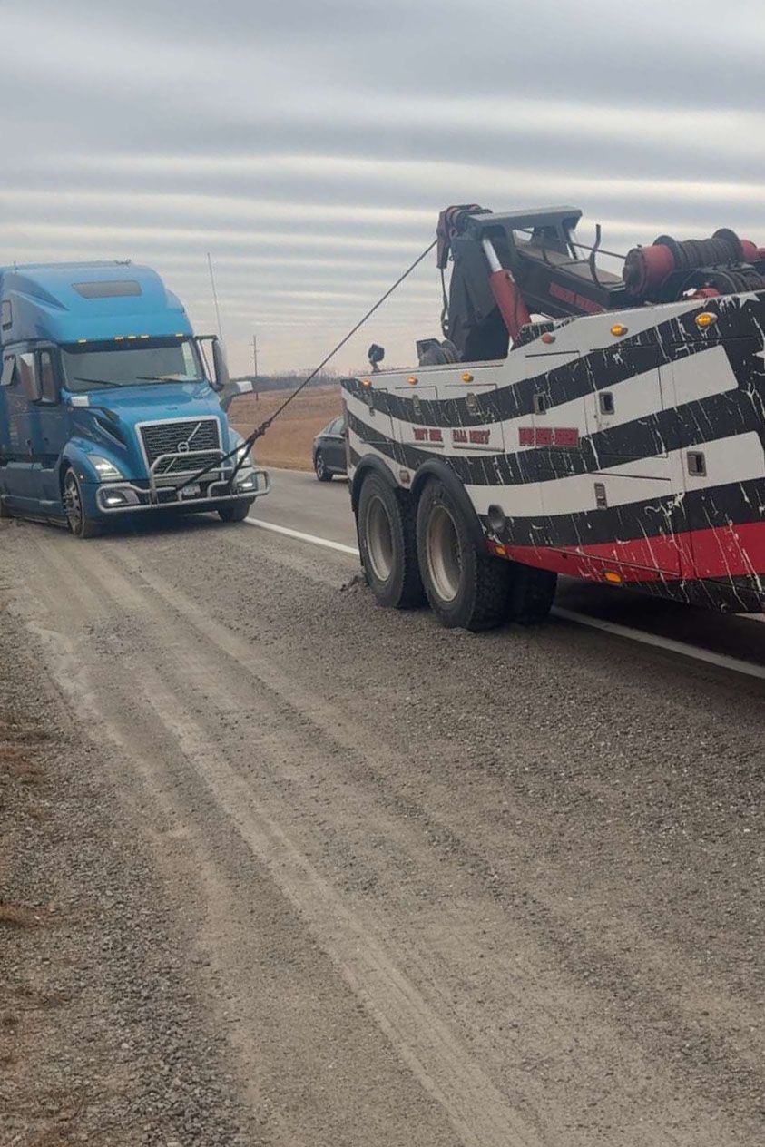A blue semi-truck being towed by a large tow truck on a gravel road, overcast sky.