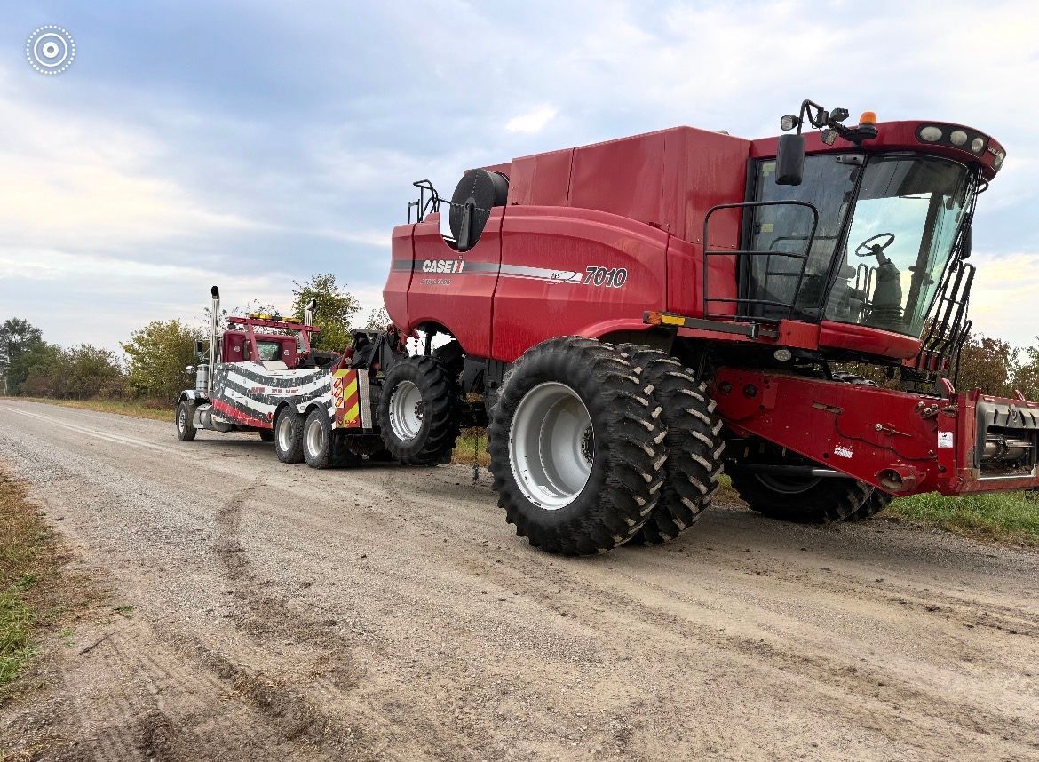 A red Case IH combine harvester being towed by a tow truck on a gravel road under a cloudy sky.