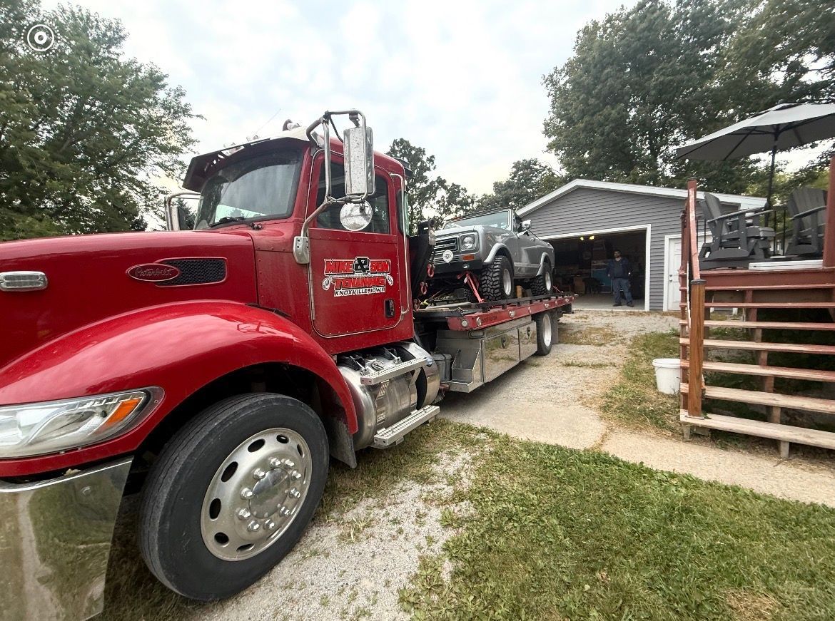 Red tow truck hauling a silver vehicle on a gravel driveway next to a garage.