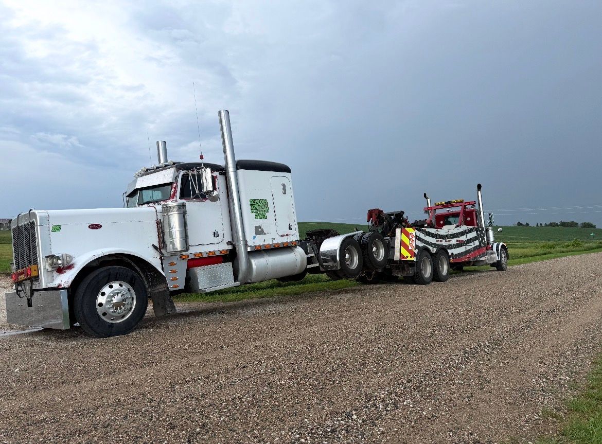 White semi-truck towing a red and black tow truck on a gravel road under a cloudy sky.