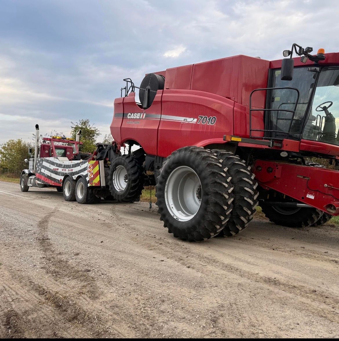 Red combine harvester being towed by a tow truck on a dirt road under a cloudy sky.