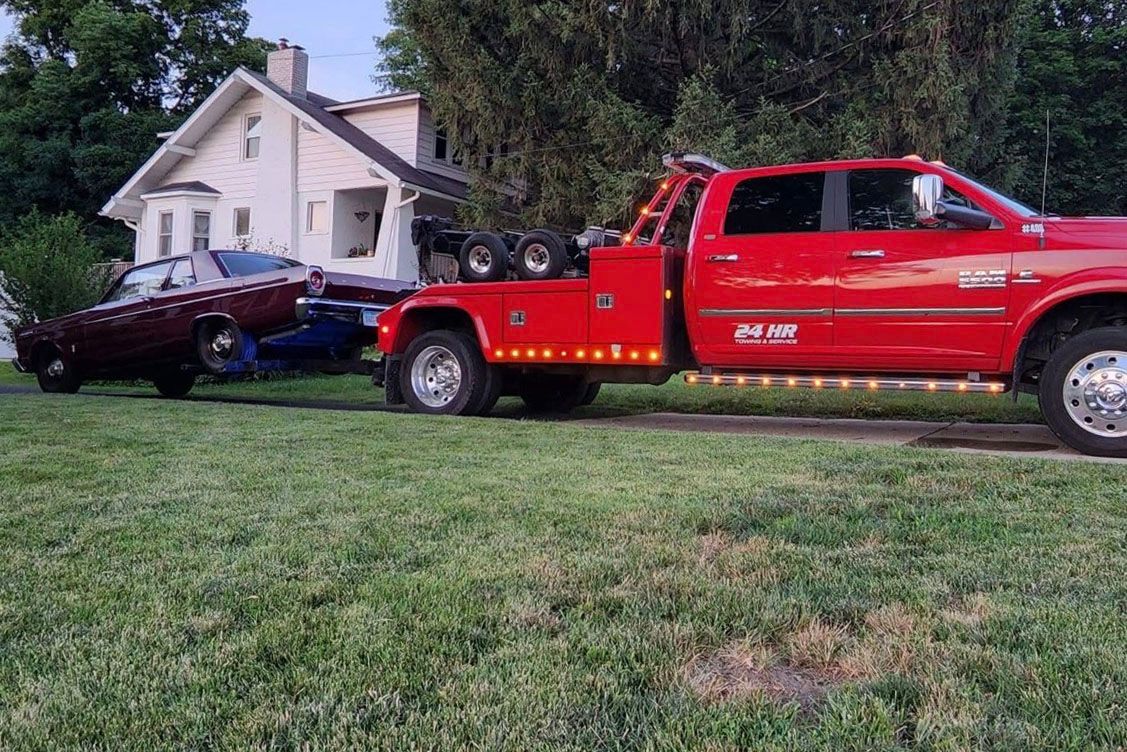 Red tow truck towing a maroon car in front of a white house on a grassy lawn.
