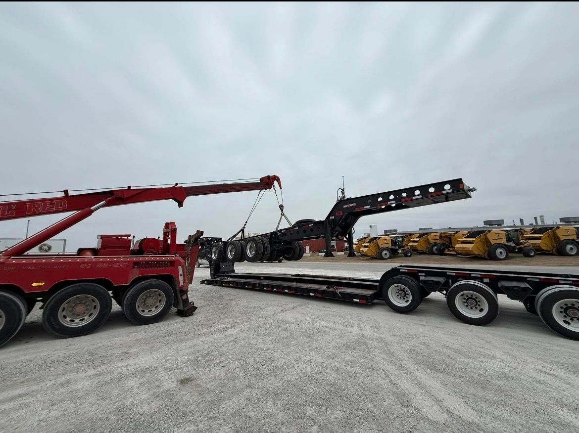 A red tow truck lifting a trailer's gooseneck. Another trailer and construction equipment are in a lot.