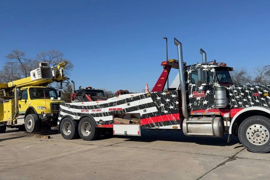 Tow truck with American flag-themed wrap towing a yellow utility truck against a blue sky.