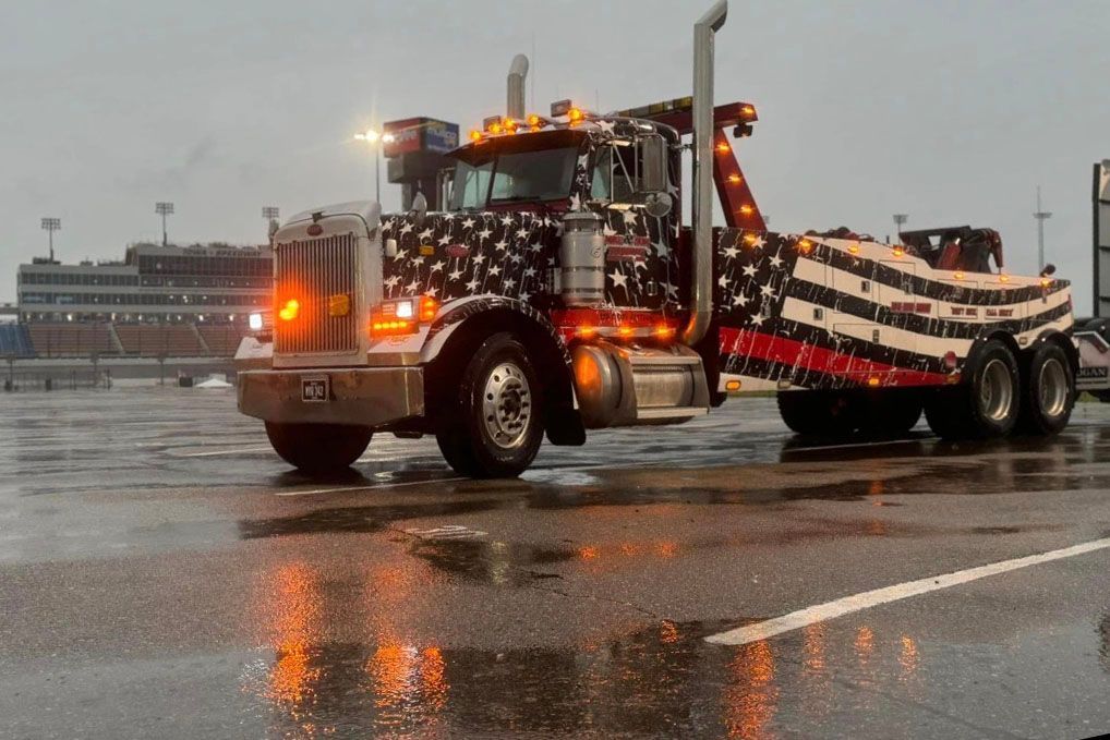 American-flag-themed tow truck parked on wet asphalt in front of a stadium on a rainy day.