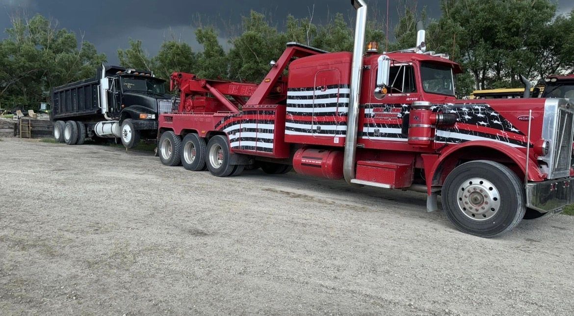Red tow truck towing a black dump truck on a gravel lot under a cloudy sky.