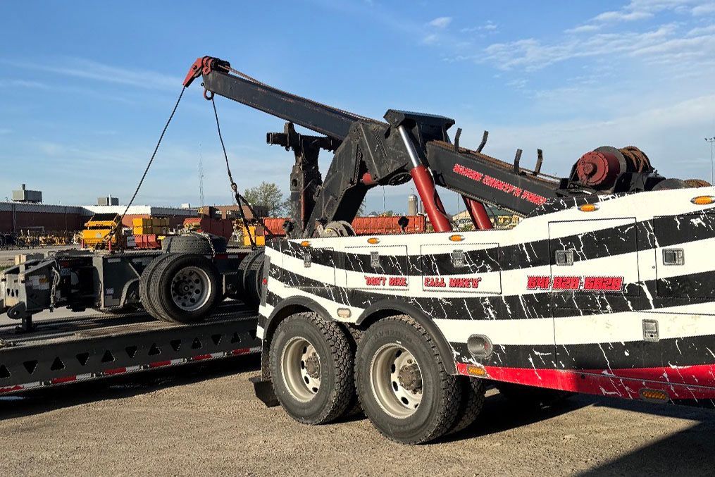 Tow truck loading a semi-truck onto a trailer. Black and white stripes on the truck, blue sky in background.