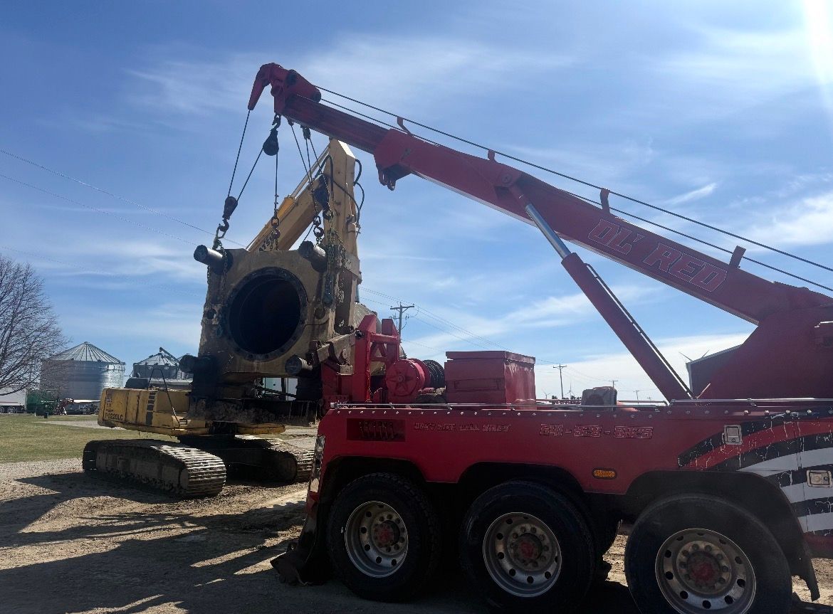Red tow truck lifting a large excavator in a field on a sunny day.