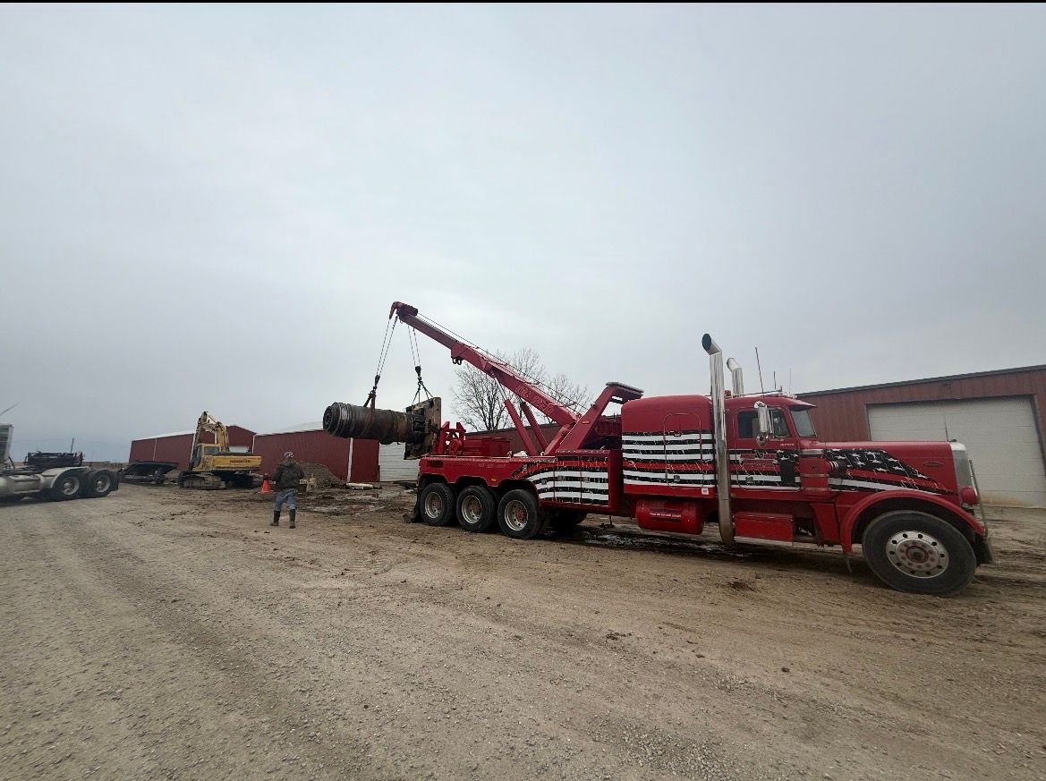 A red tow truck lifts a large object; the scene is outside near a warehouse under an overcast sky.