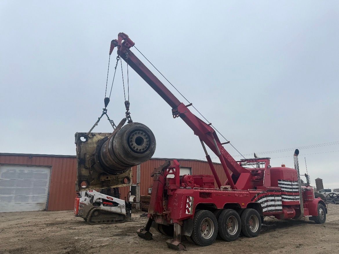 Red tow truck lifting a large machine part over a small Bobcat, cloudy day, outdoors.