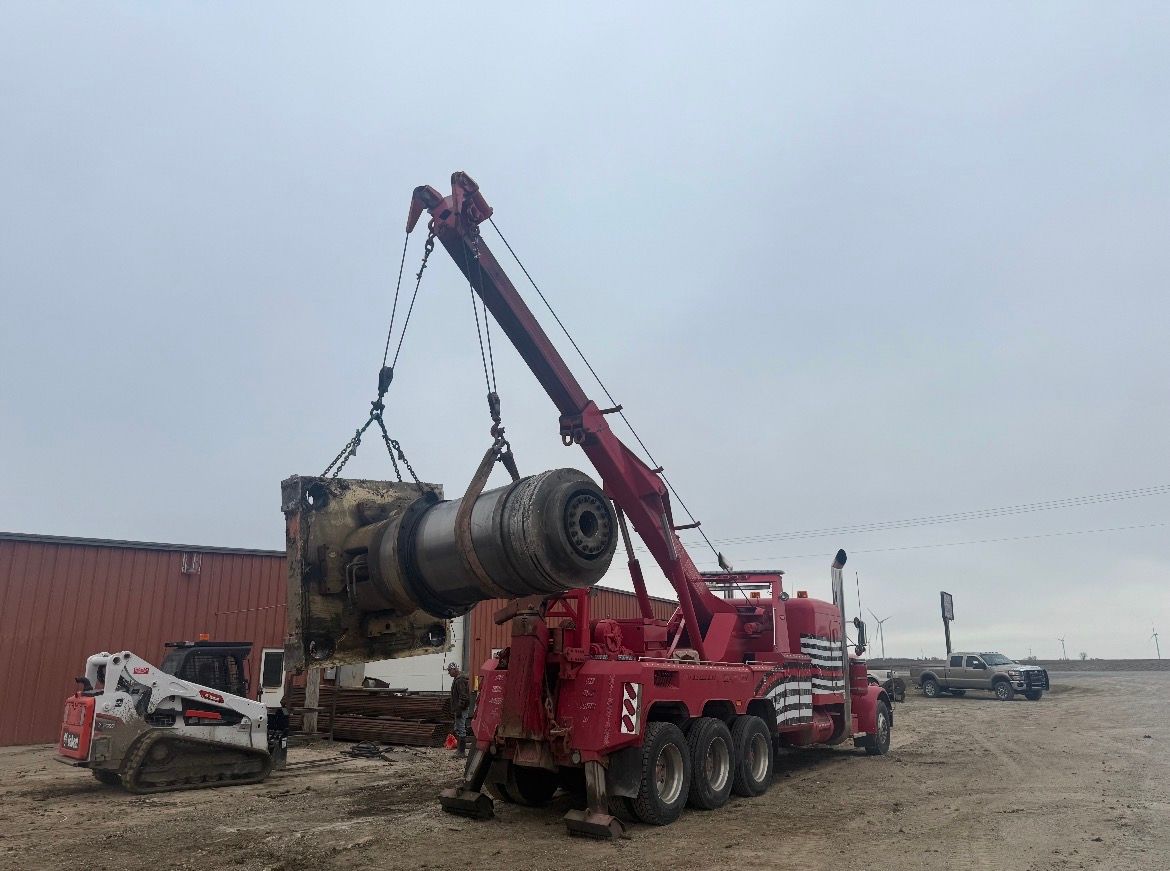 Red tow truck lifting heavy machinery on a cloudy day. A small white skid steer sits nearby.