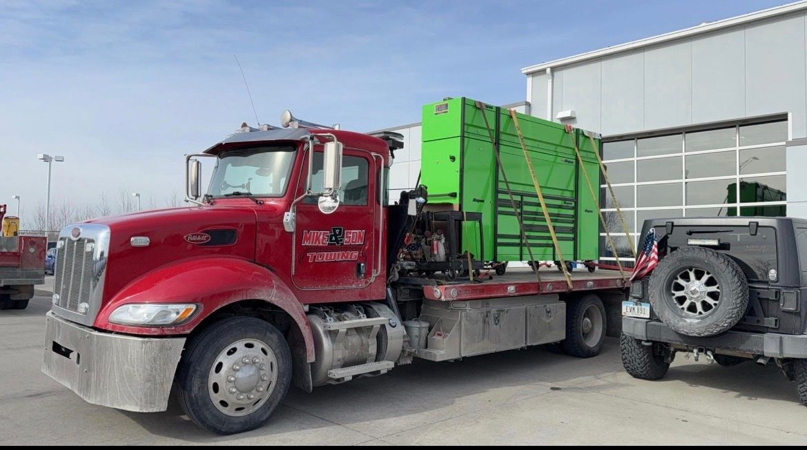 Red semi-truck with a green load on a flatbed, parked near a building with a Jeep in the side.