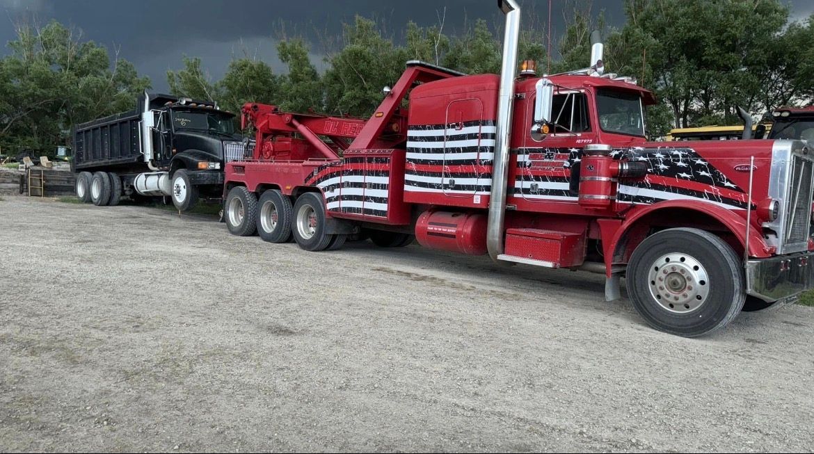 Red tow truck towing a black dump truck on a gravel lot under a cloudy sky.