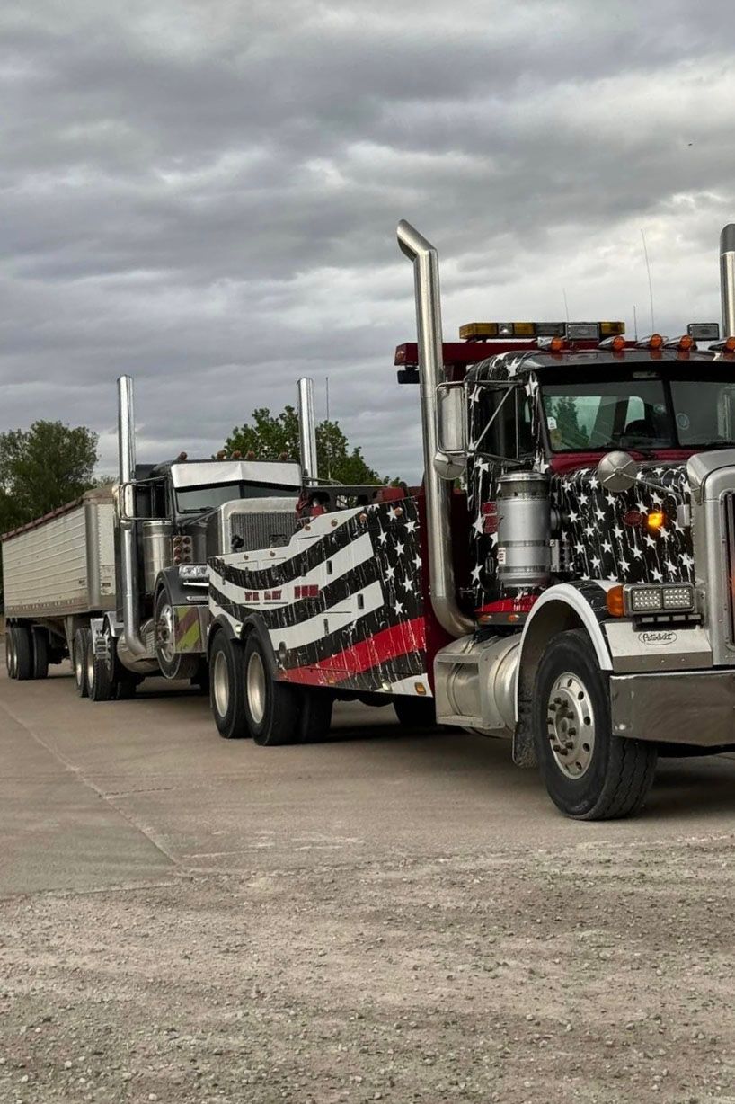 Two semi-trucks, one decorated with a patriotic design, towing another truck with a trailer on an overcast day.