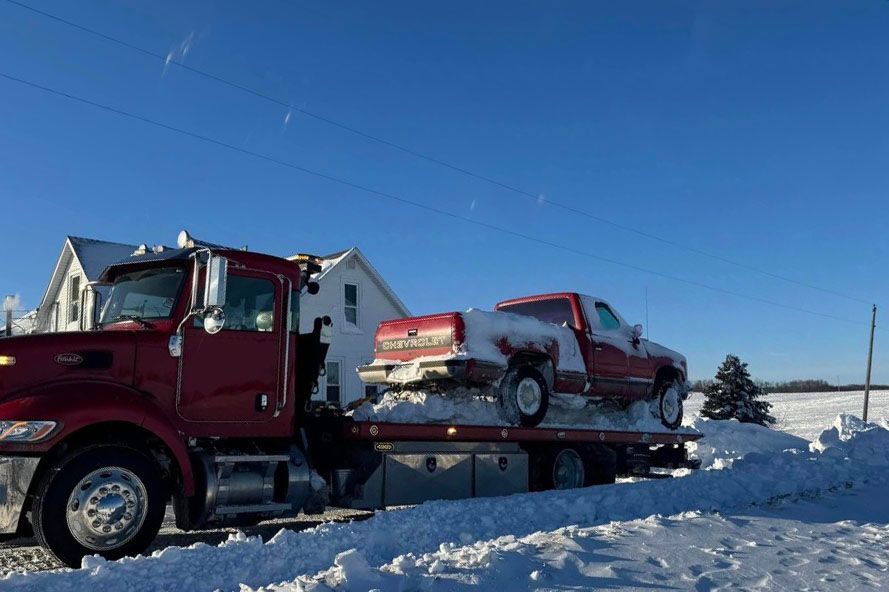 Red tow truck towing a red pickup truck on a snow-covered road near a house on a sunny day.