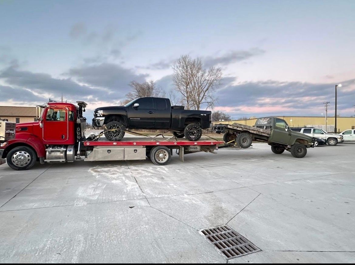 Red tow truck carrying a black pickup truck and towing a flatbed with a green vehicle. Outdoors.